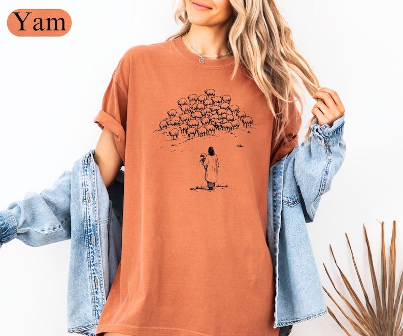 Op de afbeelding: Een terracotta-kleurig t-shirt met een zwarte illustratie van een herder en schapen. De herder houdt een lam vast en de schapen staan op een heuvel. Het woord "Yam" staat in een oranje ovaal in de linkerbovenhoek.
