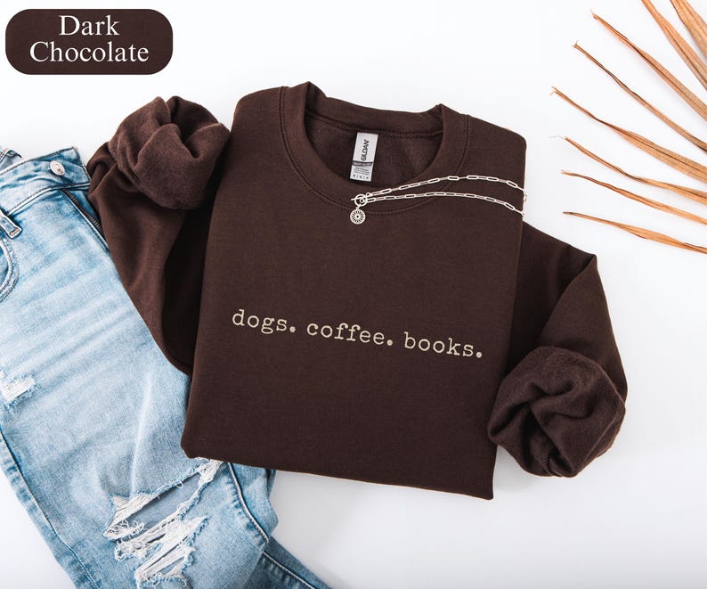 May include: Dark chocolate-colored sweatshirt with the text "dogs. coffee. books." in white. The sweatshirt is paired with light blue jeans and a silver necklace. The image also includes a dark brown label that says "Dark Chocolate".