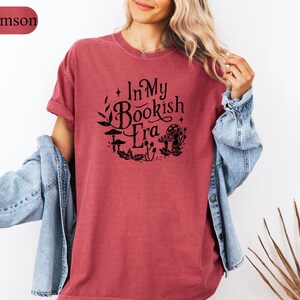 May include: Crimson-colored t-shirt with the text "In My Bookish Era" in a circular design with botanical and mushroom illustrations. The shirt is paired with a light blue denim jacket.
