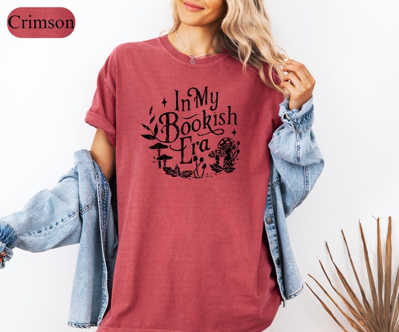 May include: Crimson-colored t-shirt with the text "In My Bookish Era" in a circular design with botanical and mushroom illustrations. The shirt is paired with a light blue denim jacket.