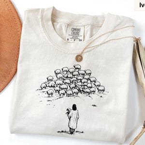 Op de afbeelding: Ivoor kleurig T-shirt met een ronde hals en korte mouwen. Het shirt heeft een zwart-wit illustratie van een persoon die een schaap vasthoudt, met een kudde schapen op een heuvel. Een gouden ketting ligt op het shirt.