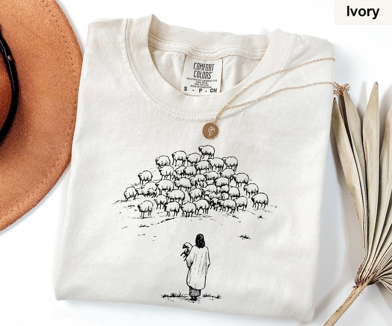 Op de afbeelding: Ivoor kleurig T-shirt met een ronde hals en korte mouwen. Het shirt heeft een zwart-wit illustratie van een persoon die een schaap vasthoudt, met een kudde schapen op een heuvel. Een gouden ketting ligt op het shirt.