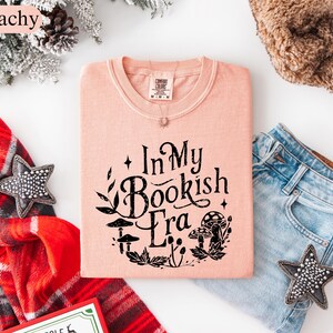 May include: A peach-colored t-shirt with the text "In My Bookish Era" in black, surrounded by botanical illustrations. The shirt is folded next to a pair of blue jeans, a star-shaped decoration, and a red plaid scarf.