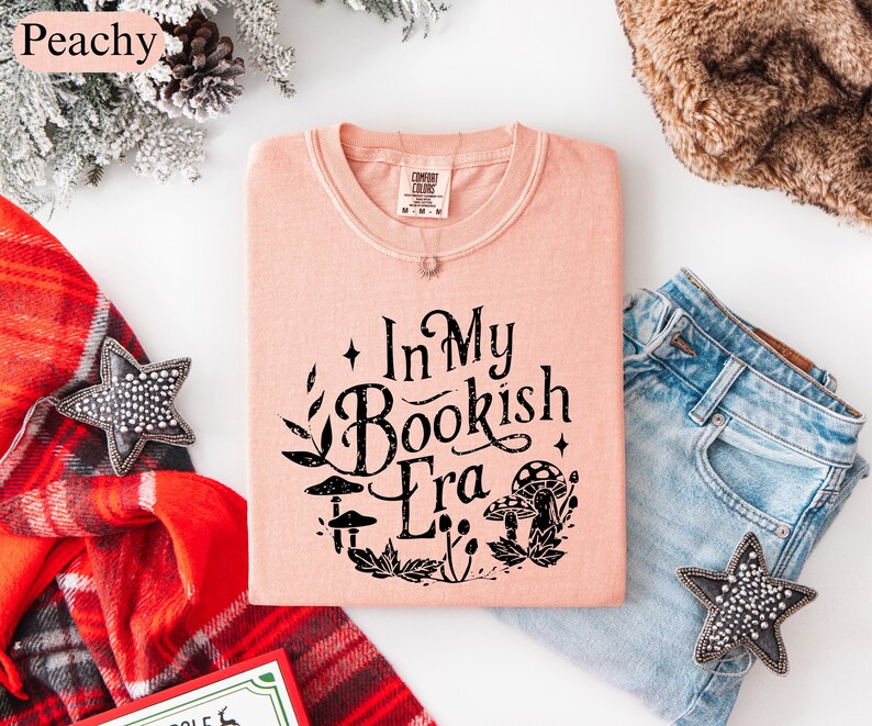 May include: A peach-colored t-shirt with the text "In My Bookish Era" in black, surrounded by botanical illustrations. The shirt is folded next to a pair of blue jeans, a star-shaped decoration, and a red plaid scarf.