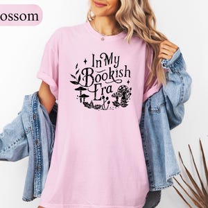 May include: A light pink t-shirt with the text "In My Bookish Era" in black, surrounded by a circular design of leaves and mushrooms. The shirt is a soft, comfortable material.