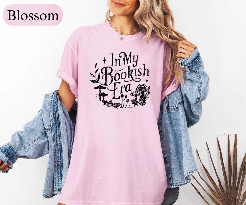 May include: A light pink t-shirt with the text "In My Bookish Era" in black, surrounded by a circular design of leaves and mushrooms. The shirt is a soft, comfortable material.