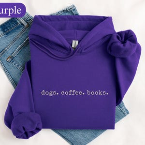 May include: A purple hooded sweatshirt with the words "dogs. coffee. books." in white. The sweatshirt is folded on top of a pair of blue jeans. The word "Purple" is in a purple oval in the upper left corner.