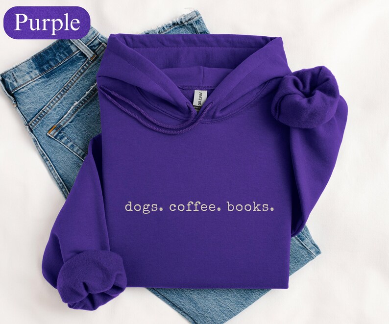 May include: A purple hooded sweatshirt with the words "dogs. coffee. books." in white. The sweatshirt is folded on top of a pair of blue jeans. The word "Purple" is in a purple oval in the upper left corner.