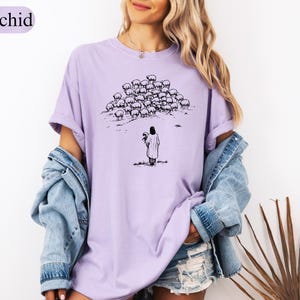 Op de afbeelding: Orchidee-kleurig T-shirt met een zwart-wit illustratie van een persoon voor een kudde schapen. De persoon houdt een haan vast. Het shirt heeft een relaxte pasvorm en korte mouwen.