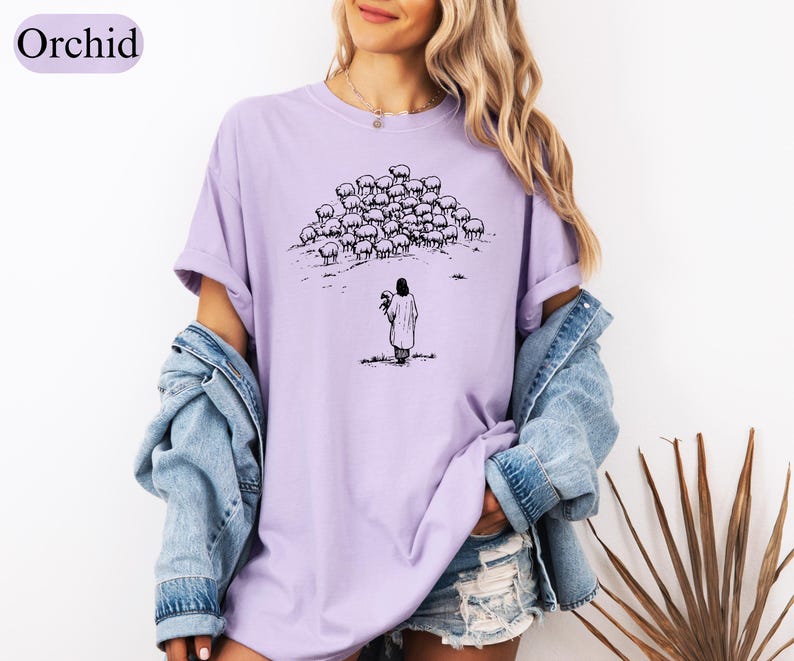 Op de afbeelding: Orchidee-kleurig T-shirt met een zwart-wit illustratie van een persoon voor een kudde schapen. De persoon houdt een haan vast. Het shirt heeft een relaxte pasvorm en korte mouwen.