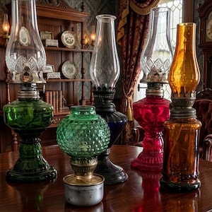 May include: A collection of antique oil lamps in various colors, including green, red, amber, and blue. The lamps have glass chimneys and ornate metal bases. The lamps are arranged on a dark wooden table, with a vintage interior in the background.