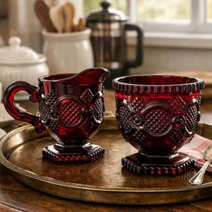 May include: A ruby red glass creamer and sugar bowl set with ornate detailing. The creamer has a handle and spout, while the sugar bowl is open. Both pieces sit on a decorative metal tray, showcasing a vintage aesthetic.