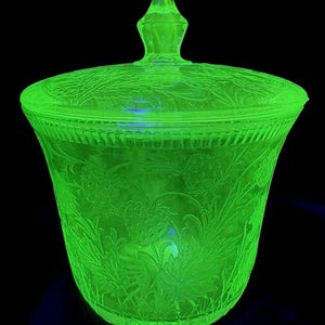 May include: A glowing green glass jar with a lid, showcasing intricate floral and leaf patterns. The jar has a flared shape and a decorative finial on the lid. The glass appears to be vintage, with a vibrant, almost neon, green hue.