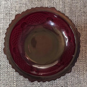 May include: A ruby red glass bowl with a scalloped edge. The bowl has a textured pattern on the sides and a smooth, dark center. The rim of the bowl has a silver-colored trim. The bowl is sitting on a gray and white patterned surface.