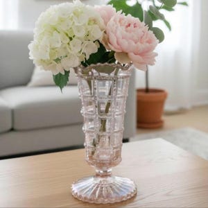 May include: A clear, pink-tinted glass vase with a grid pattern and scalloped base holds white hydrangeas and a pink peony. The vase sits on a wooden table, showcasing a classic floral arrangement.