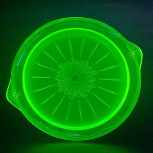 May include: A round, glowing green glass dish with a floral design in the center. The dish has a raised rim and two small handles. The design features a central flower with radiating lines.
