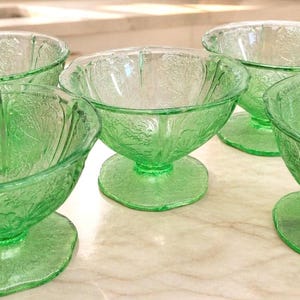 May include: A set of five green glass dessert bowls. Each bowl has a pedestal base and a scalloped rim. The glass has a floral pattern. The bowls are arranged on a light-colored surface.