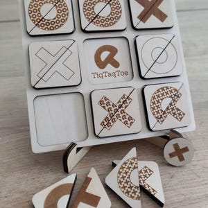 Op de afbeelding: Houten Tic-Tac-Toe spel met een lichte houten afwerking. Het spelbord heeft negen vierkanten met gegraveerde X'en en O's, sommige met patronen. De tekst "TiqTaqToe" is zichtbaar op het bord. Inclusief houten speelstukken.