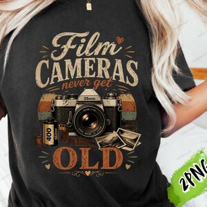 Op de afbeelding: Zwart T-shirt met een vintage camera-ontwerp en de tekst "Film Cameras Never Get Old". Het ontwerp bevat een camera, filmrol en foto's, in een retro kleurenschema van bruin, oranje en beige. Digitaal product.