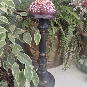 May include: A decorative mushroom ornament with a dark brown cap and white spots, set on a black, ornate stand. The stand has a round base and a fluted column. The background includes green plants and foliage.