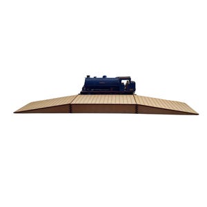 May include: A dark blue model train locomotive sits on a tan, brick-effect platform. The platform has a sloped ramp on one side. The model train is a detailed replica of a classic steam engine.