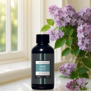 May include: A black bottle of "Pure Lilac" reed diffuser fragrance oil. The label features the brand name "Aronica" and a teal and black design. The bottle is next to a vase of purple lilacs, with a window in the background.