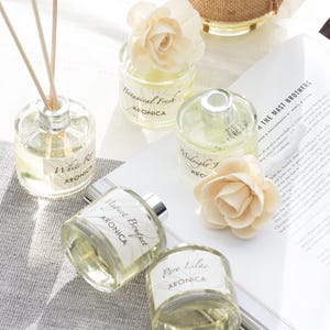 May include: An assortment of glass aromatherapy diffusers with floral accents and wooden reeds. The diffusers are labeled with various scents, including "White Rose," "Perfect Bouquet," and "Pure Lilac" by ARONICA. The image also includes a book and decorative flowers.