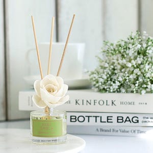 May include: A clear glass diffuser bottle with a white flower and wooden reeds. The bottle has a green label that reads "MINI SHOLA". The diffuser sits on a white marble surface, with books and a bouquet of white flowers in the background.