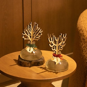 May include: Two decorative glass bottles with wooden tree-like structures on top, sitting on a round wooden table. One bottle rests on a wooden base, while the other has a red ribbon. The bottles have gold accents and are in a warm-toned room.