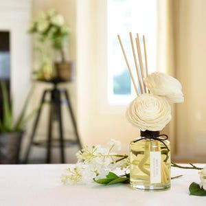 May include: A clear glass diffuser bottle filled with a yellow liquid, featuring wooden reeds and decorative white flower accents. The bottle is tied with a brown string and has a label that reads "White Rose".