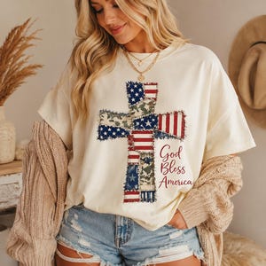 May include: Cream-colored t-shirt featuring a cross design made of American flag and camouflage patterns. The text "God Bless America" is printed in a script font. The shirt is worn with denim shorts.