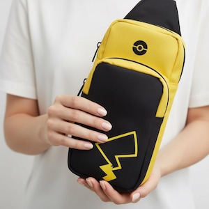 Könnte beinhalten: Gelb-schwarze Umhängetasche mit Pikachu-Design. Die Tasche hat einen gelben oberen Bereich mit einem schwarzen Pokéball-Symbol und einen schwarzen unteren Bereich mit einer gelben Blitzgrafik. Die Tasche wird von einer Person gehalten.