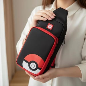 May include: Black and red crossbody bag with a graphic of a red, white, and black ball. The bag has a zippered main compartment and a small red square patch with a white and red graphic. The bag is held by a person.