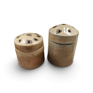 Pode incluir: Dois potes cilíndricos de cerâmica com tampas, apresentando um esmalte bege liso e brilhante no topo e um acabamento castanho terroso e texturizado na base. Cada tampa é abobadada e perfurada com vários pequenos orifícios circulares.