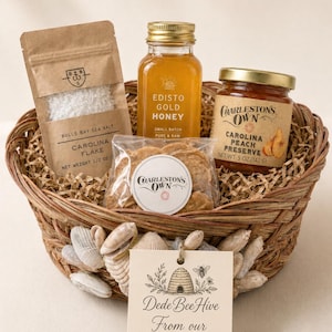 May include: A woven basket filled with a pouch of Bulls Bay Sea Salt, a glass bottle of Edisto Gold Honey, a jar of Charleston's Own Carolina Peach Preserve, and a bag of cookies. A tag reads Dede Bee Hive From our hive to yours.