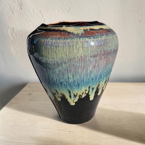 May include: A ceramic vase with a wide, rounded body and a narrow opening. The glaze features a layered effect, transitioning from a deep black base at the bottom to a vibrant blend of turquoise, blue, and earthy brown tones dripping downwards.