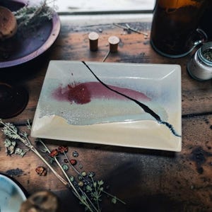 May include: A rectangular ceramic tray with a glossy finish, featuring a cream-colored base, a horizontal band of muted blue, and a prominent, textured deep red circular accent. A single, thin, dark, jagged line cuts diagonally across the surface.