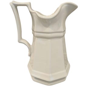 May include: A cream-coloured ceramic jug featuring a hexagonal body, a wide pouring spout, and a tall, angular handle. The vessel has a smooth, glossy finish with defined vertical ridges along the sides and a flared base for stability.
