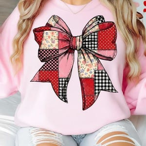 May include: A light pink sweatshirt featuring a large, patchwork bow design. The bow incorporates various patterns, including red polka dots, black and white gingham, and floral prints, with a central pink section. The bow is a prominent graphic on the front of the shirt.
