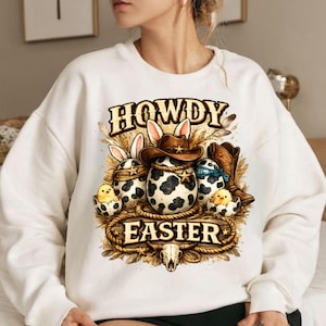 May include: White sweatshirt with a graphic featuring Easter eggs with cow print, cowboy hats, and bunny ears. The text reads "HOWDY EASTER" in a Western-style font, with a skull at the bottom. The design includes a rope border and feathers.