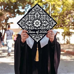 Może przedstawiać: Czarna czapka absolwenta z białym kwiatowym wzorem i napisem "Class of 2026". Czapka ma złoty chwost i jest trzymana przed osobą w todze.