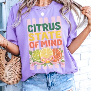 Könnte beinhalten: Ein hellviolettes T-Shirt mit einem Grafikdruck und dem Text CITRUS STATE OF MIND in Grün, Gelb und Orange. Das Design zeigt Illustrationen einer Limette, einer Zitrone und einer Orangenscheibe, die mit einem rosa Band und kleinen Blumen verziert sind.