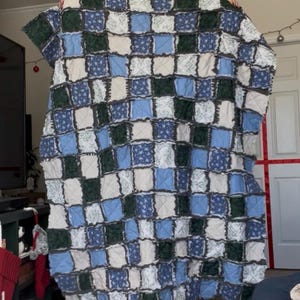 May include: A handmade patchwork quilt featuring a grid pattern of squares in shades of blue, green, and white. The quilt has a textured, rag-style finish, and is likely made of soft fabric.