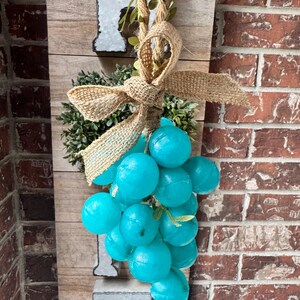 May include: A rustic wooden sign featuring a cluster of turquoise grapes, a burlap bow, and greenery. The sign has metal letter accents and is suspended by a rope. The sign is a decorative item.