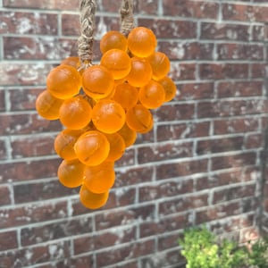 May include: A cluster of orange, spherical beads, resembling grapes, is suspended by a thick, braided rope. The beads have a translucent quality, and the rope is a natural, light brown color. The background is a brick wall.