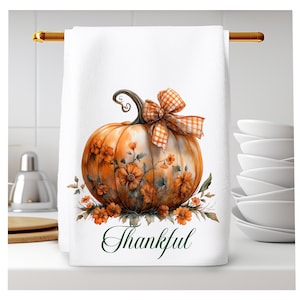Op de afbeelding: Een witte keukendoek met een aquarel-illustratie van een oranje pompoen met een geruite oranje en witte strik. De pompoen is omringd door oranje bloemen en de tekst "Thankful" is in groene cursieve letters onder de pompoen geschreven.