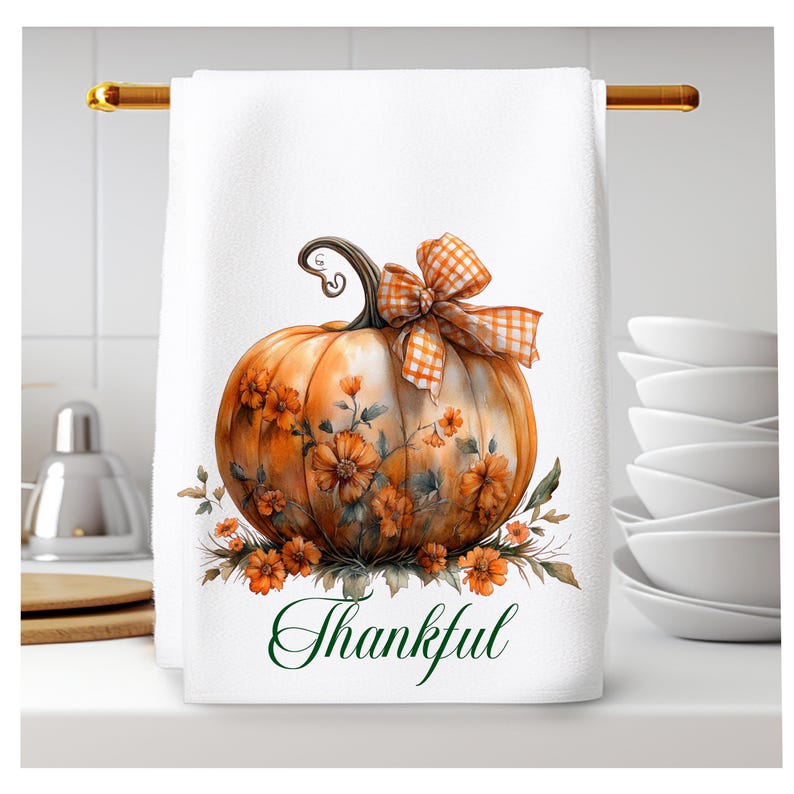 Op de afbeelding: Een witte keukendoek met een aquarel-illustratie van een oranje pompoen met een geruite oranje en witte strik. De pompoen is omringd door oranje bloemen en de tekst "Thankful" is in groene cursieve letters onder de pompoen geschreven.