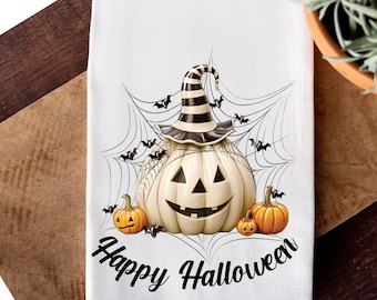 May include: White kitchen towel with a black and white pumpkin wearing a witch's hat. The pumpkin is surrounded by a spider web with bats flying around it. The text "Happy Halloween" is written below the pumpkin.