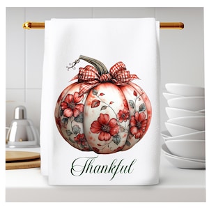 Op de afbeelding: Een witte keukendoek met een rode en witte geruite strik op een pompoen met rode bloemen en groene bladeren. De tekst "Thankful" is in groen onder de pompoen gedrukt.