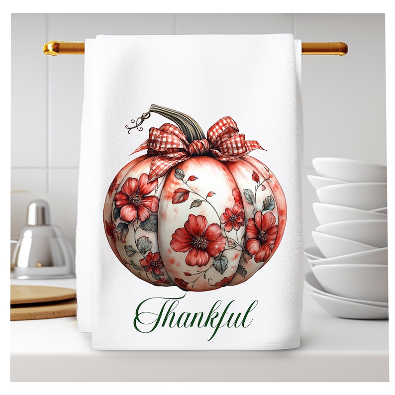 Op de afbeelding: Een witte keukendoek met een rode en witte geruite strik op een pompoen met rode bloemen en groene bladeren. De tekst "Thankful" is in groen onder de pompoen gedrukt.
